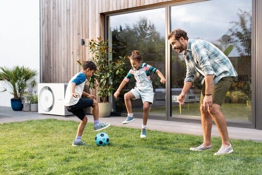Vater und zwei Kinder spielen Fußball im Garten. Im Hintergrund sieht man am Haus eine Wärmepumpe.
