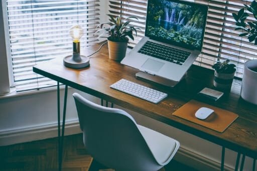 Ein Laptop auf Laptop-Ständer, davor eine Tastatur auf einem aufgeräumten Schreibtisch. Homeoffice vor heruntergelassenen Jalousien.