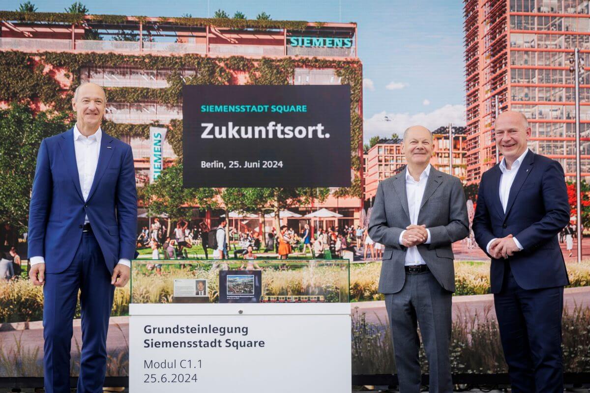 Ein Gruppenbild vom Vorsitzenden der Siemens AG Roland Busch, Bundeskanzler Olaf Scholz und Berlins Bürgermeister Kai Wegner bei der Grundsteinlegung für Siemensstadt Square. Der Hintergrund (eine Visualisierung) zeigt moderne, begrünte Gebäude des neuen Stadtteils. Vor der Leinwand steht ein Tisch mit der Beschriftung "Grundsteinlegung Siemensstadt Square".