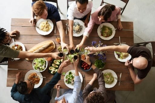 Menschen sitzen gemeinsam am Esstisch und stoßen beim gemeinsamen Essen an. Feste wie diese können zu einer guten Nachbarschaft beitragen.
