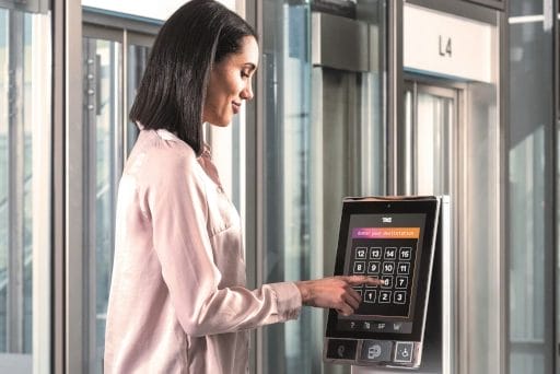 Eine Frau steht seitlich zum Aufzug im 4. Stockwerk und drückt auf einem Touchpad die Taste 6.