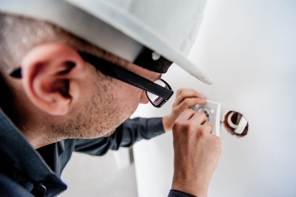 Elektriker mit Helm und Brille leicht nach vorne gebeugt vor einer Wand und hält eine Steckdosenabdeckung in der Hand.