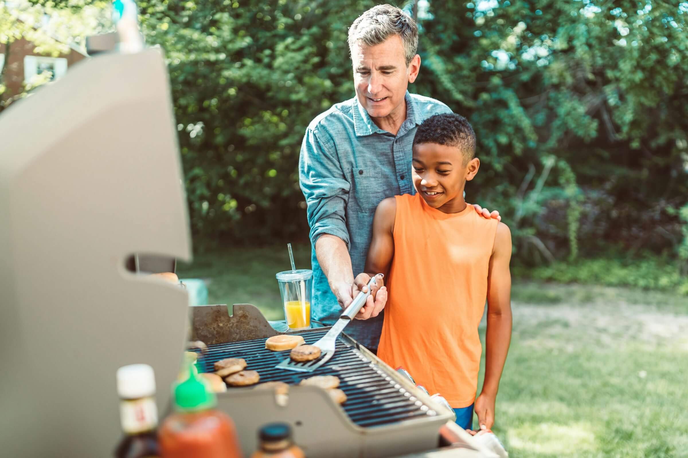 Ein Mann bringt einem Jungen am Gartengrill bei, wie er Frikadellen grillen kann.