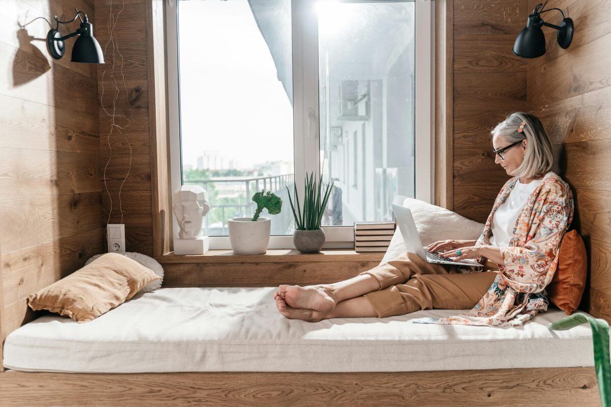 Eine Frau sitzt auf dem Bett eines Apartments und schaut etwas in ihrem Laptop nach. Im Hintergrund ist die Wohnanlage der Ferienimmobilie zu sehen.