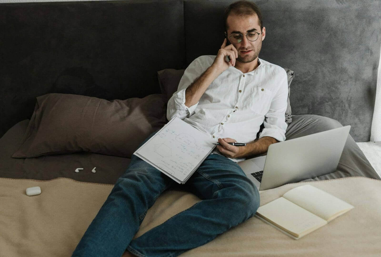Mann sitzt entspannt auf einem Bett, spricht am Telefon, arbeitet mit einem Laptop und hat ein Notizbuch aufgeschlagen – ein Beispiel für Homeoffice im Schlafzimmer.