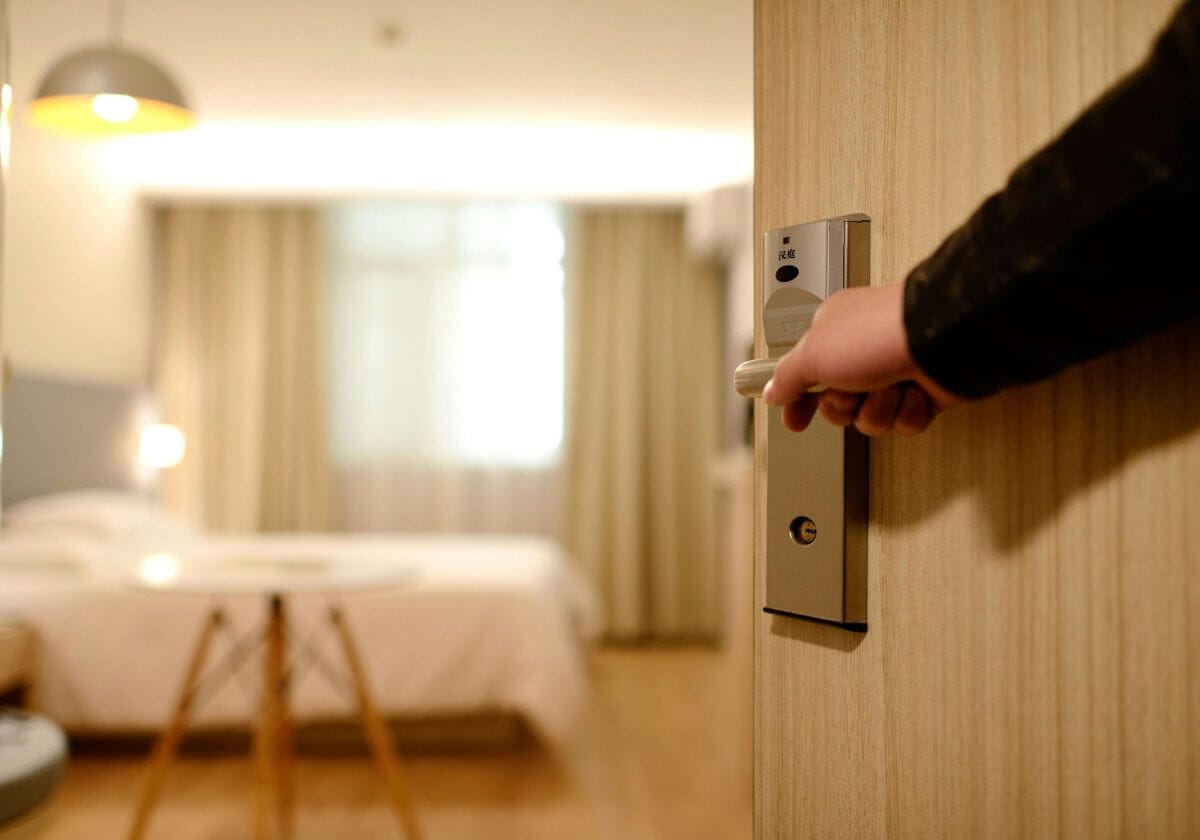 Eine Hand öffnet die Tür zu einem möblierten Hotelzimmer.