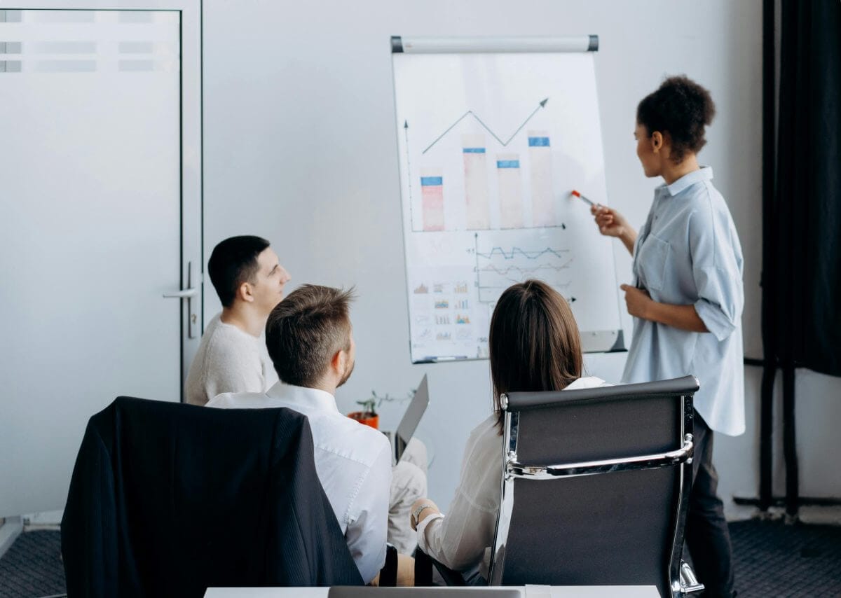 Eine Frau präsentiert Diagramme auf einem Flipchart vor einer Gruppe von drei Personen in einem Meeting. Die Grafik zeigt steigende Kurven, die Fortschritt und erfolgreiche Planung symbolisieren, passend zum Thema Quartiersmanagement.