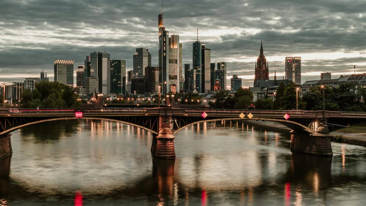 Blick von der Ignatz-Bubis-Brücke auf Frankfurts Skyline bei Abenddämmerung: Eine Brücke mit beleuchteten Markierungen spannt sich über den Main, dessen ruhige Wasseroberfläche die Lichter der Stadt spiegelt. Im Hintergrund ragen moderne Wolkenkratzer neben historischen Gebäuden in den Himmel.