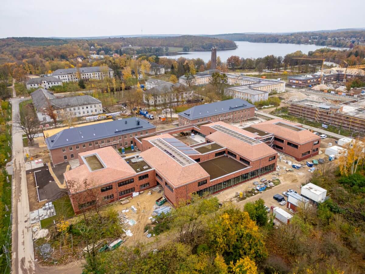 Luftaufnahme des neu gebauten Schulcampus im Quartier Krampnitz, umgeben von alten Gebäuden und einer Baustelle, mit See im Hintergrund.