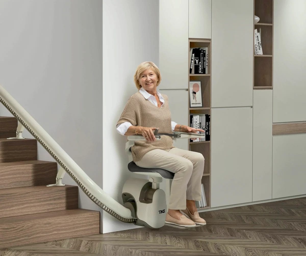 Eine Frau mittleren Alters mit blonden Haaren sitzt auf einem Treppenlift am unteren Ende der Treppe. Ihr Blick ist in die Kamera gerichtet.